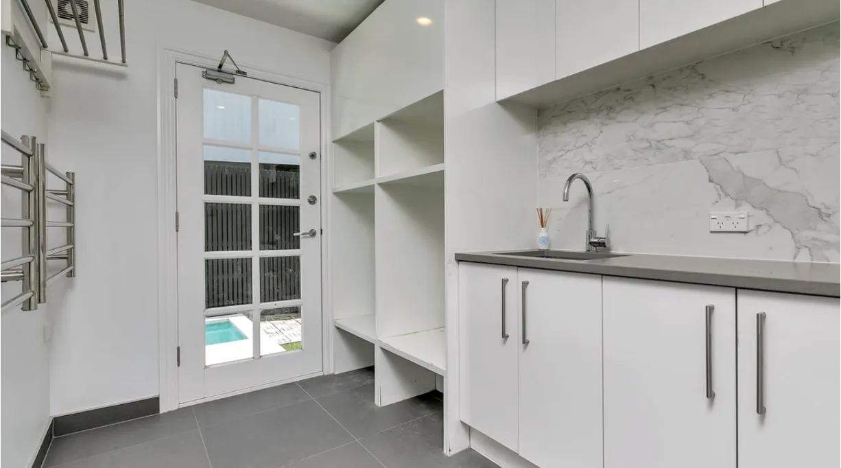 A white laundry with the washing machine and dryer set on a raised plinth, open shelving above, a stone benchtop alongside and a glazed door to the garden with the pool beyond.