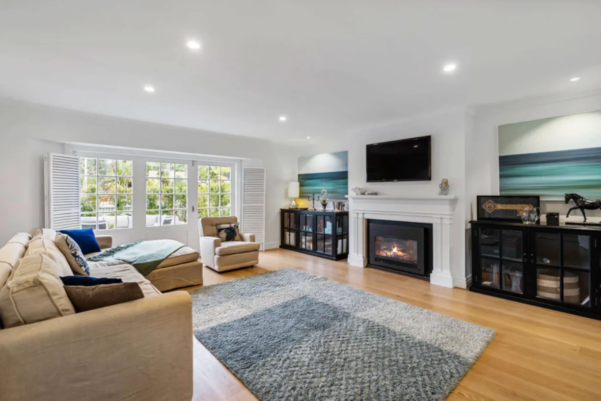 A living room with a gas fire under a white mantel, plantation shutters at the windows and framed artwork above the hearth.