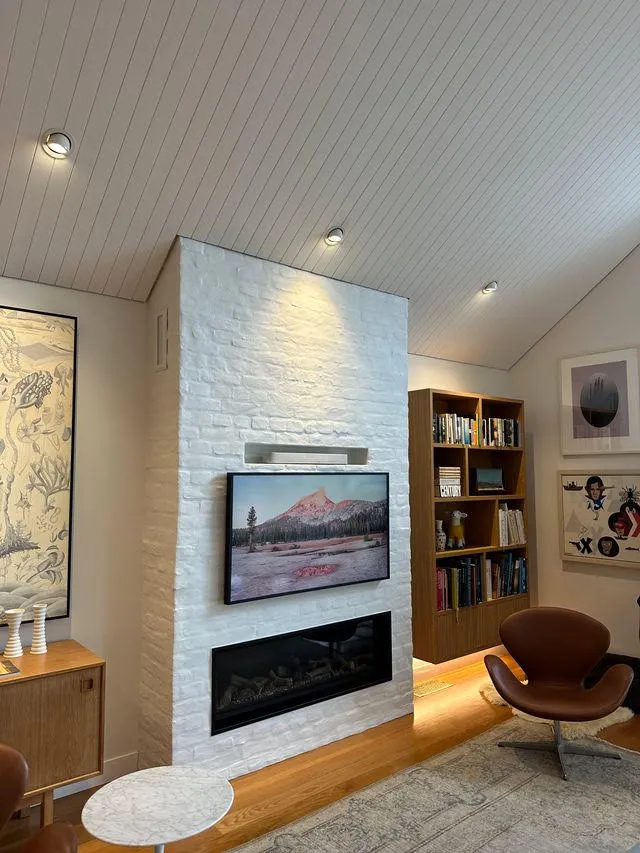 A living room with a white brick fireplace and inset fire, timber bookshelves alongside and a tan leather chair by the hearth.