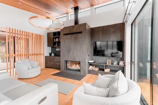 A living room under a cedar-lined raked ceiling, with a dark veined stone fireplace surround, a circular timber pendant and clerestory glazing above.