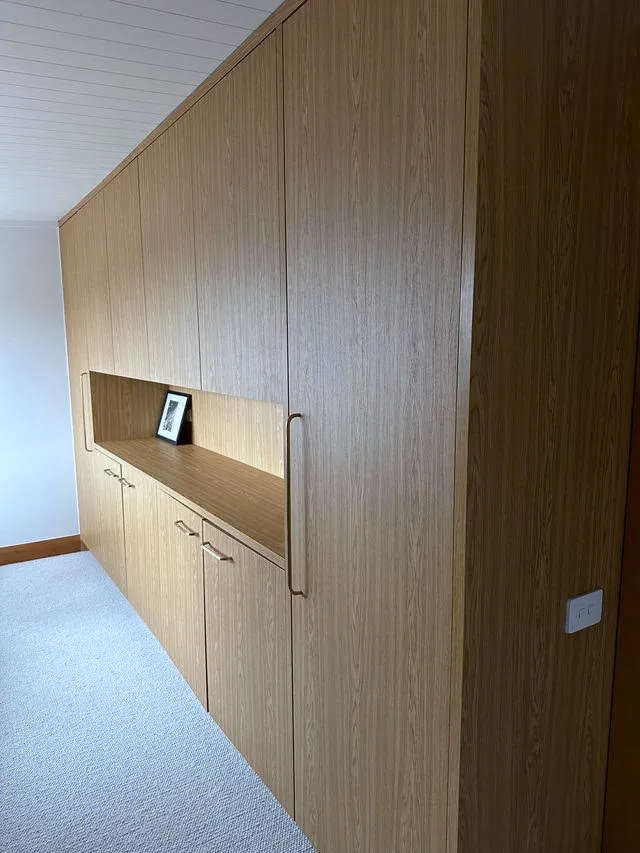 An oak joinery wall in a bedroom with a long recessed shelf, concealing a door through to the wardrobe behind it.