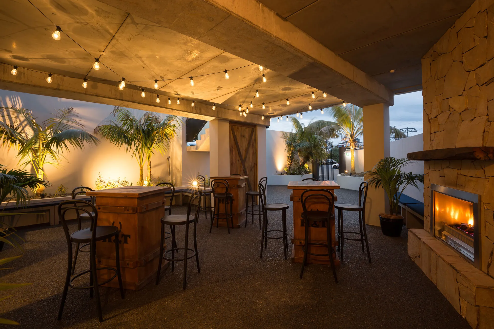 A covered outdoor bar at dusk, with festoon lighting overhead, barrel tables and stools, a stone fireplace alight and palms along the edges.