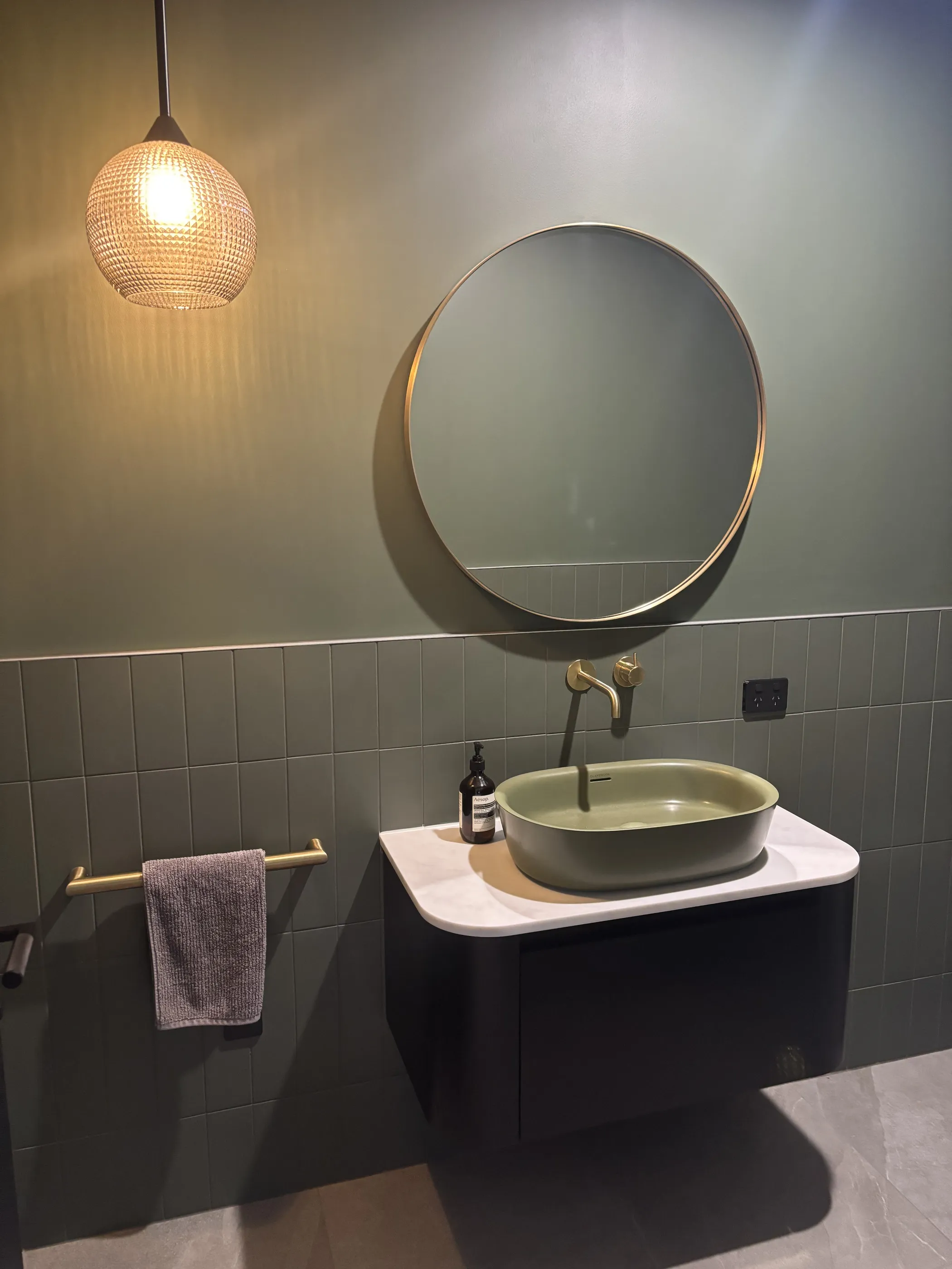 A powder room with sage-green stacked tile, an olive-green vessel basin on a black vanity, brass tapware and a round brass-rimmed mirror below a rattan globe pendant.