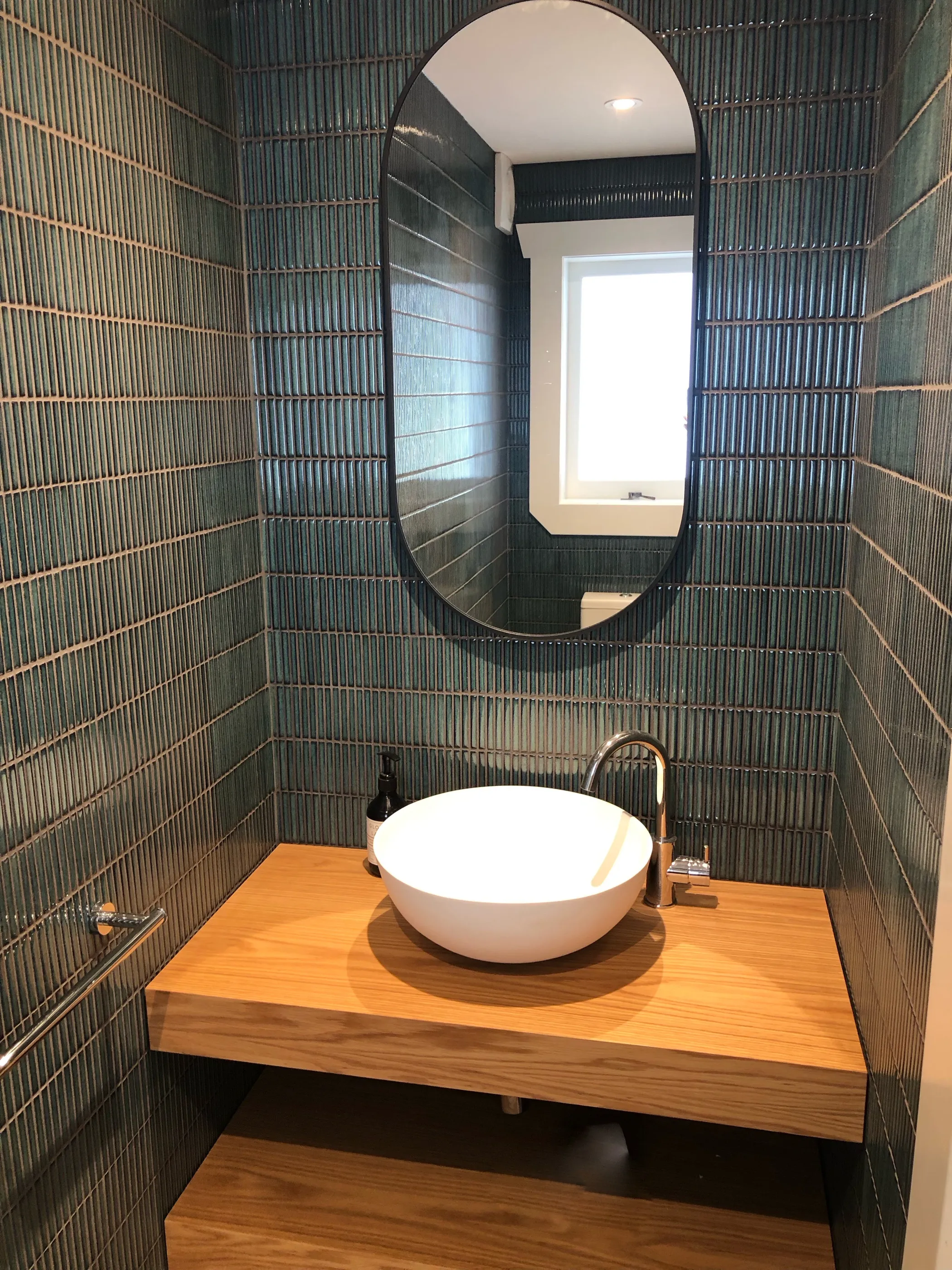 A powder room lined in deep teal fluted tile, with an oval mirror, a brass tap and a white basin on a floating timber vanity.