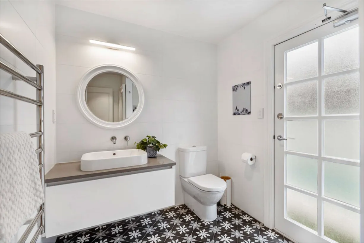 A powder room with patterned black and white encaustic floor tiles, a round white mirror and wall-mounted tapware over a white basin.