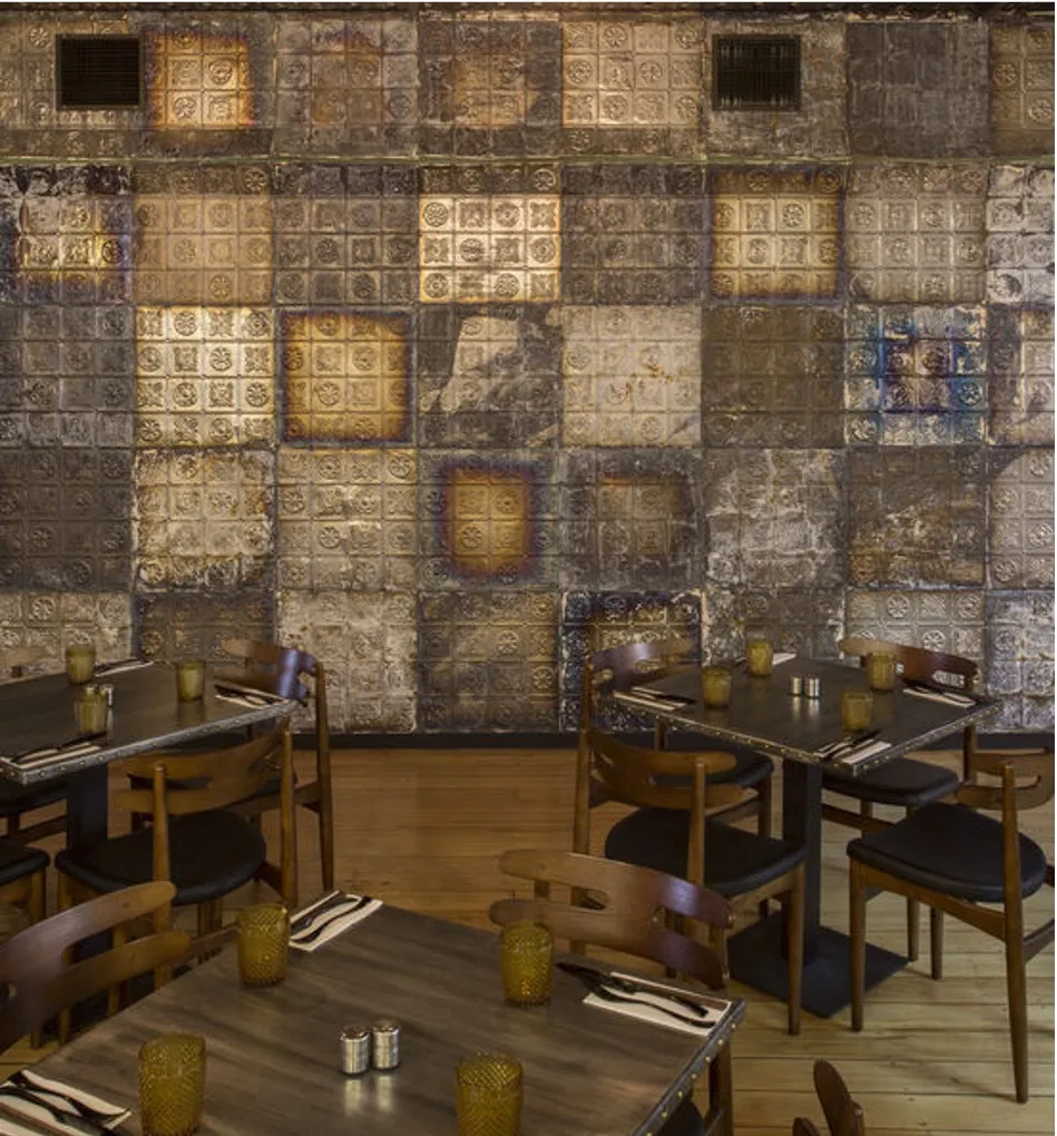 A feature wall of patchworked pressed tin panels, warmly uplit, with timber dining settings and candles in the foreground.