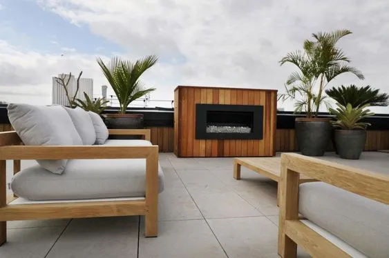 A rooftop terrace with teak seating, a timber-clad outdoor fireplace and potted palms, open to the city skyline.