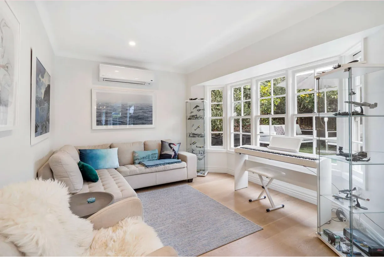 A softer sitting room with a bay window, an upright piano against the wall and armchairs drawn around a low table.