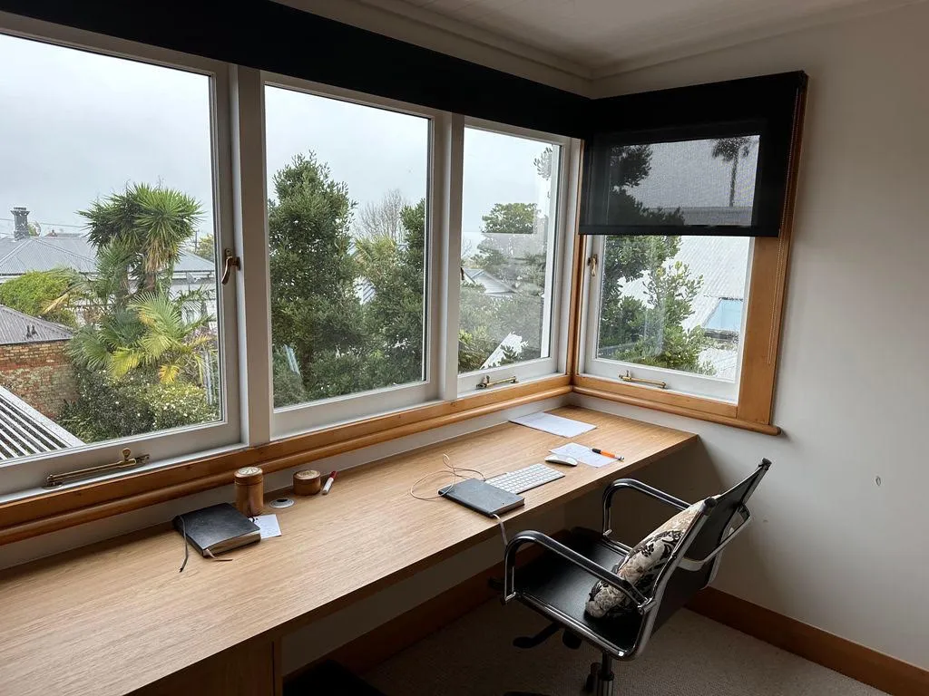 A study with a timber desk running the length of a bay of windows, an office chair drawn up to it and treetops beyond.