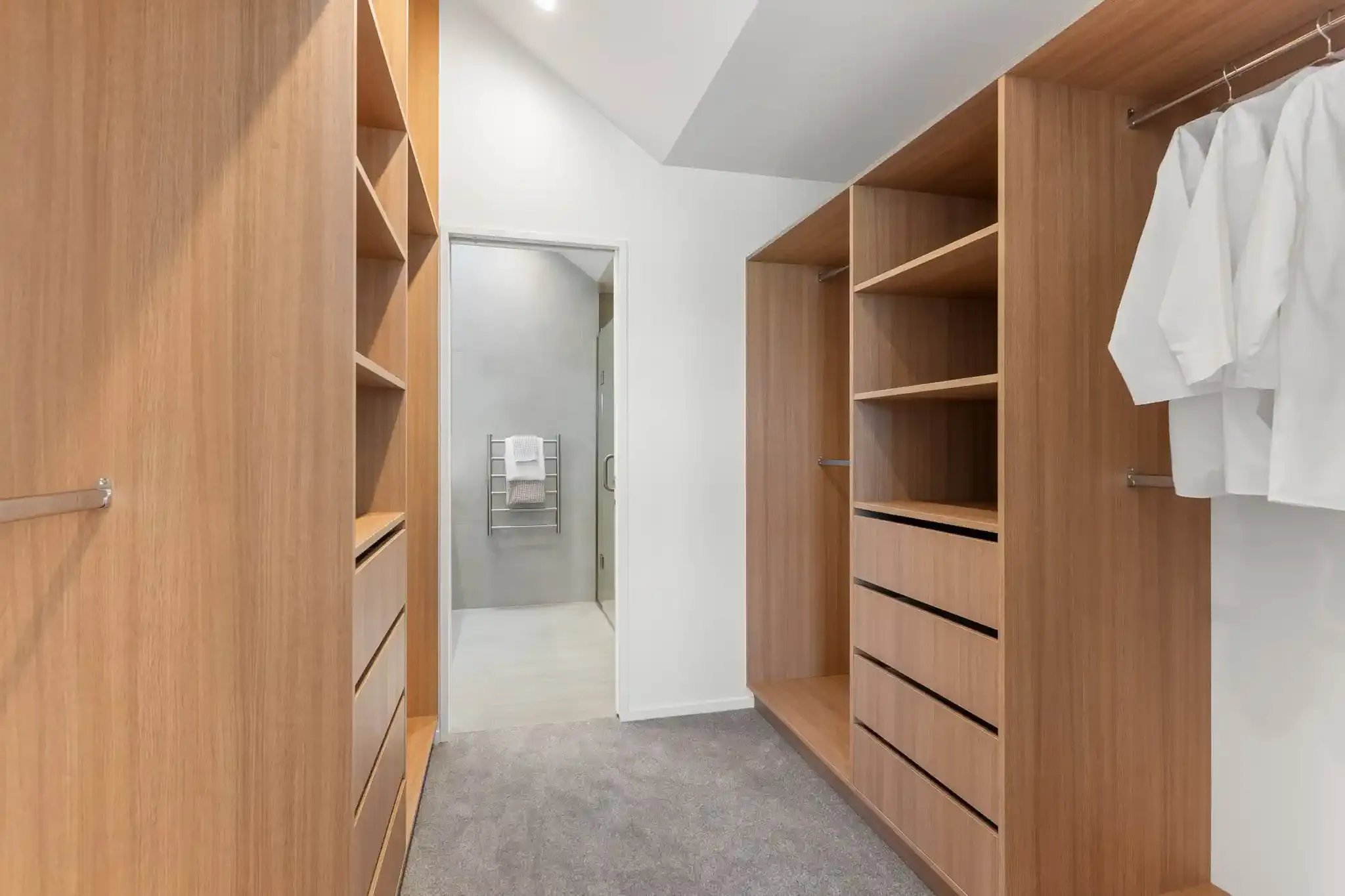 A walk-in wardrobe fitted out in oak, with open shelving, drawers and hanging rails, and an ensuite visible through the doorway.
