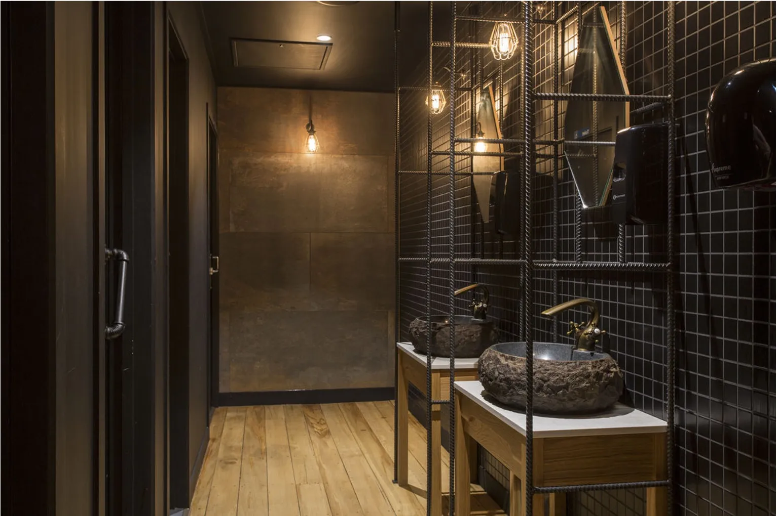 A washroom screened in rebar, with stone vessel basins, brass tapware, black square mosaic tiling and Edison pendants overhead.
